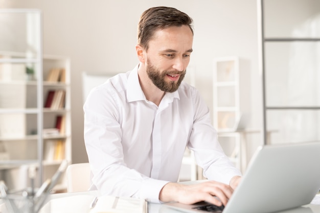 Jeune analyste en chemise blanche en tapant sur le clavier d'un ordinateur portable tout en saisissant des informations ou en naviguant sur le net