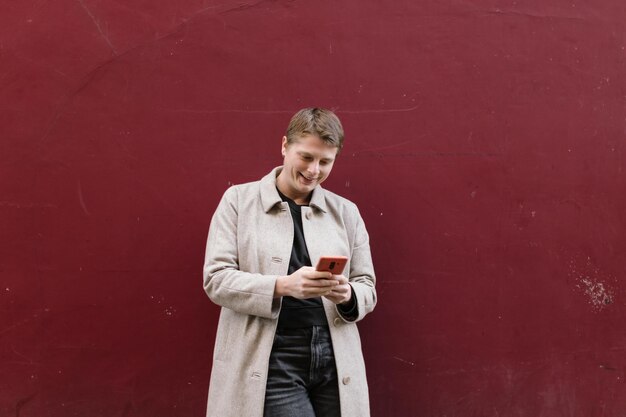 Jeune androgyne à la mode souriant utilisant un téléphone portable allongé contre un mur rouge