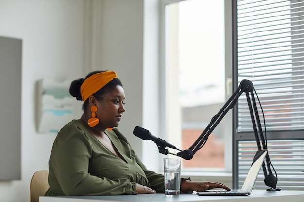 Jeune animateur de radio assis à la table devant un ordinateur portable et parlant au microphone travaillant à la radio