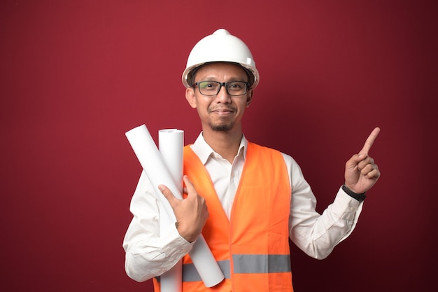 Jeune architecte asiatique portant un casque, tenant un plan papier du projet présentant quelque chose de son côté.