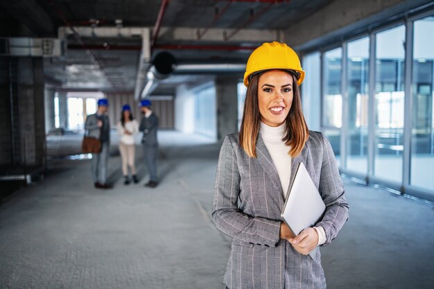 Jeune architecte caucasienne attrayante réussie avec casque sur la tête debout à l'intérieur du bâtiment en processus de construction et tenant un ordinateur portable dans les mains.