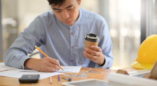 Un jeune architecte est en train de rédiger un design de maison et tient une tasse de café dans un bureau moderne.