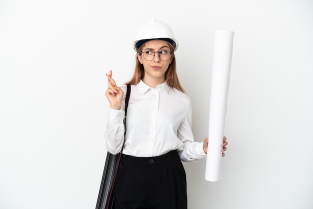 Jeune architecte femme avec casque et tenant des plans isolés sur un mur blanc avec les doigts qui se croisent et souhaitant le meilleur