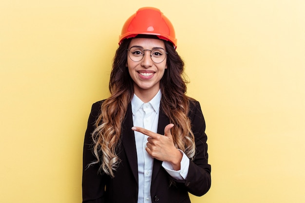 Jeune architecte femme métisse isolée sur fond jaune souriant et pointant de côté, montrant quelque chose dans un espace vide.