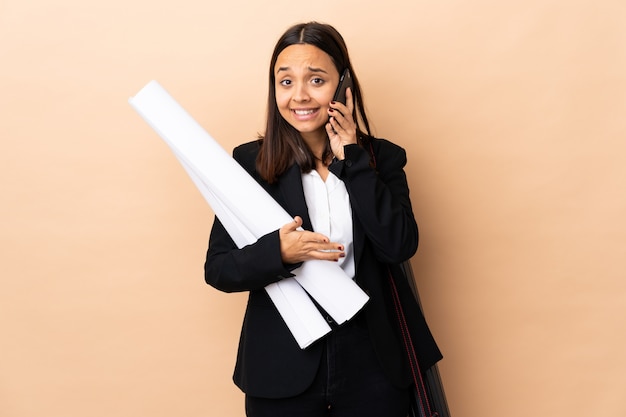 Jeune architecte femme tenant des plans sur le mur en gardant une conversation avec le téléphone mobile avec quelqu'un