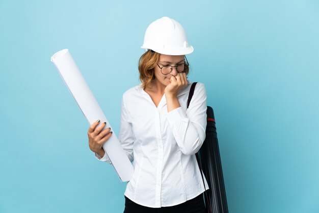 Jeune architecte géorgien femme avec casque et tenant des plans sur fond isolé ayant des doutes