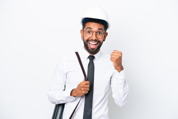 Jeune architecte homme brésilien avec casque et tenant des plans isolés sur fond blanc célébrant une victoire en position de vainqueur