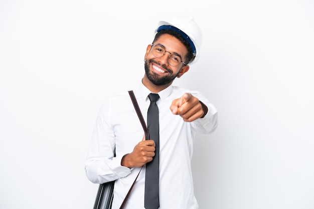 Jeune architecte homme brésilien avec casque et tenant des plans isolés sur fond blanc pointant vers l'avant avec une expression heureuse