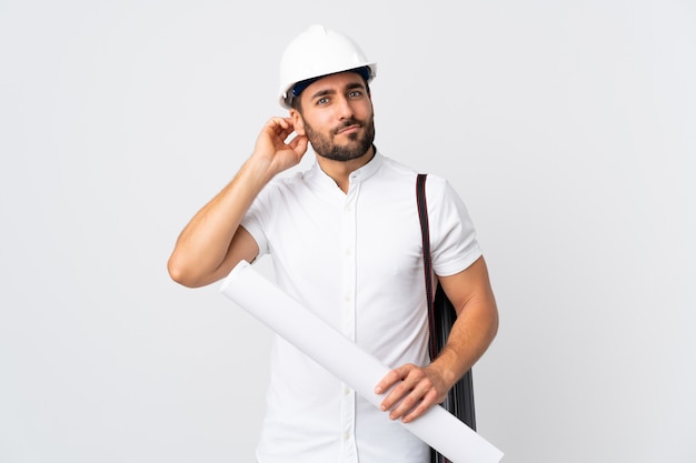 Jeune architecte homme avec casque et tenant des plans isolé sur mur blanc ayant des doutes