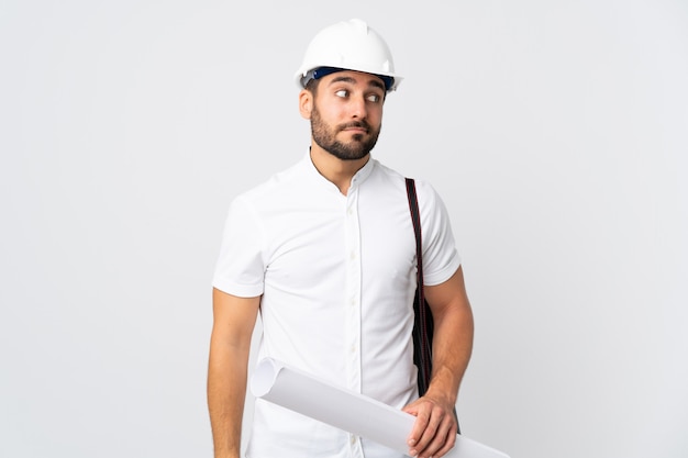 Jeune architecte homme avec casque et tenant des plans isolé sur mur blanc faisant des doutes geste regardant côté