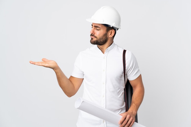 Jeune architecte homme avec casque et tenant des plans isolé sur mur blanc tenant copyspace avec des doutes