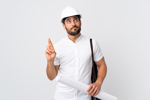 Jeune architecte homme avec casque et tenant des plans isolés sur mur blanc avec les doigts traversant et souhaitant le meilleur