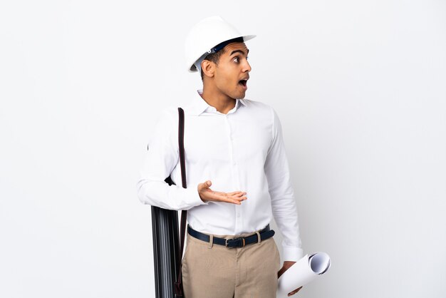 Jeune architecte homme avec casque et tenant des plans sur un mur blanc isolé _ avec une expression de surprise tout en regardant côté