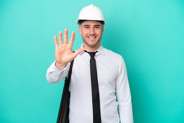 Jeune architecte homme caucasien avec casque et tenant des plans isolés sur fond bleu comptant cinq avec les doigts