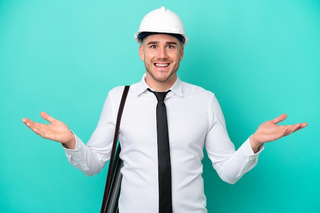 Jeune architecte homme caucasien avec casque et tenant des plans isolés sur fond bleu avec une expression faciale choquée