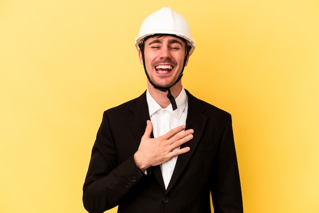 Jeune architecte homme caucasien isolé sur fond jaune rit fort en gardant la main sur la poitrine.