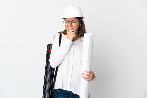 Jeune architecte lituanien femme avec casque et tenant des plans isolés sur un mur blanc à la recherche sur le côté et souriant