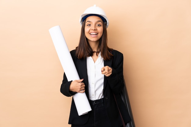 Jeune architecte woman holding blueprints sur isolé surpris et pointant vers l'avant