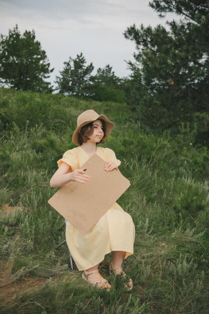 jeune artiste de 1013 ans vêtue d'une robe jaune et d'un chapeau de paille dans la nature peignant un tableau