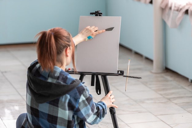 Une jeune artiste adolescente avec un couteau à palette dans les mains s'apprête à peindre une peinture à l'huile. Une toile blanche avec copie est située sur le chevalet noir.