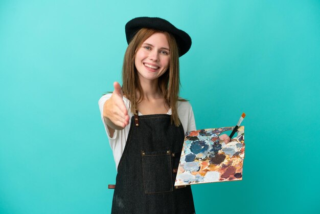 Jeune artiste anglais femme tenant une palette isolée sur fond bleu se serrant la main pour conclure une bonne affaire