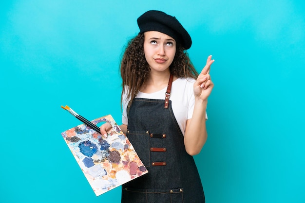 Jeune artiste arabe femme tenant une palette isolée sur fond bleu avec les doigts croisés et souhaitant le meilleur
