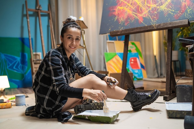Un jeune artiste est assis sur un sol sécurisé avec du carton parmi des peintures et des peintures tenant un pinceau