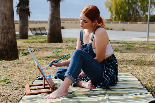 Jeune artiste féminine rousse caucasienne peint une toile de chevalet dans le parc à l'extérieur