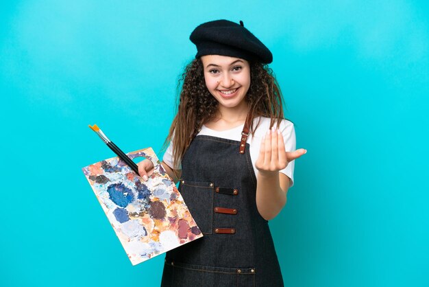 Jeune artiste femme arabe tenant une palette isolée sur fond bleu invitant à venir avec la main Heureux que vous soyez venu