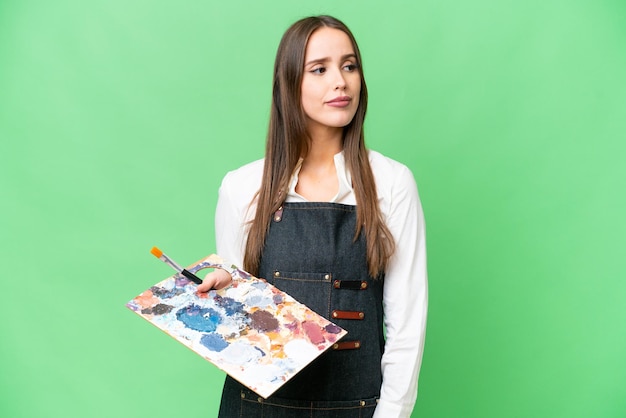 Jeune artiste femme tenant une palette sur fond isolé chroma key regardant sur le côté