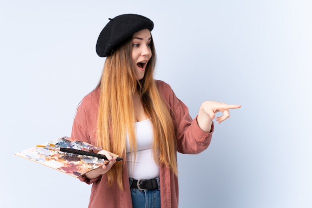 Photo jeune artiste femme tenant une palette isolée sur le mur bleu, pointant le doigt sur le côté