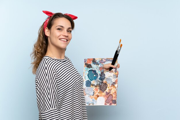 Jeune artiste femme tenant une palette sur un mur bleu isolé souriant beaucoup