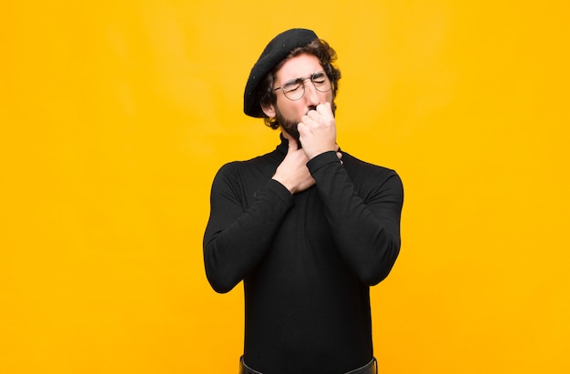 Photo jeune artiste français homme se sentant mal avec un mal de gorge et des symptômes de la grippe, toussant avec la bouche couverte contre le mur orange