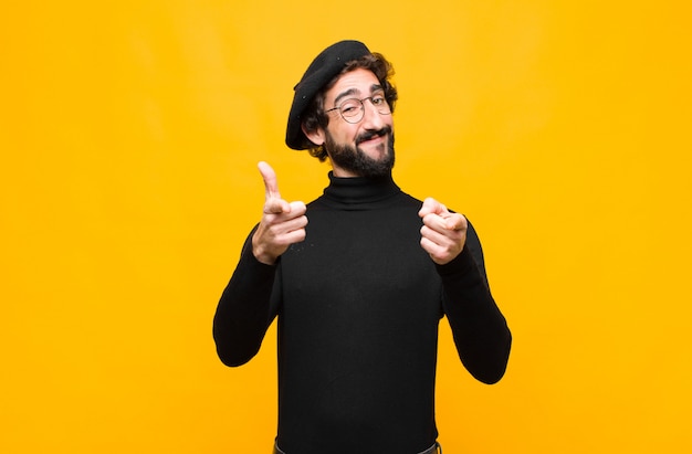 Jeune artiste français homme souriant avec une attitude positive, réussie et heureuse pointant vers la caméra, faisant signe de pistolet avec les mains contre le mur orange