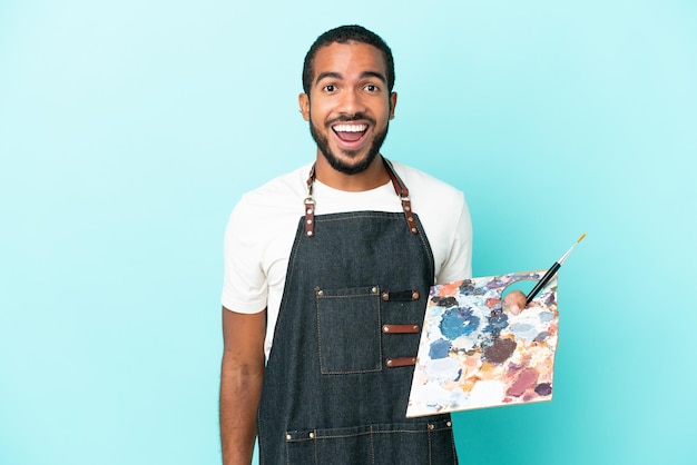 Jeune artiste homme latin tenant une palette isolée sur fond bleu avec une expression faciale surprise