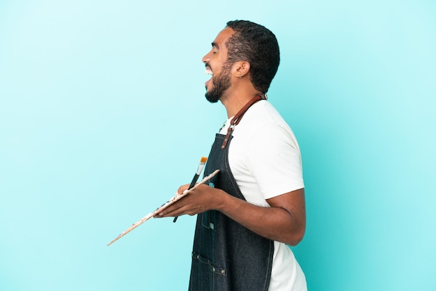 Photo jeune artiste homme latin tenant une palette isolée sur fond bleu riant en position latérale