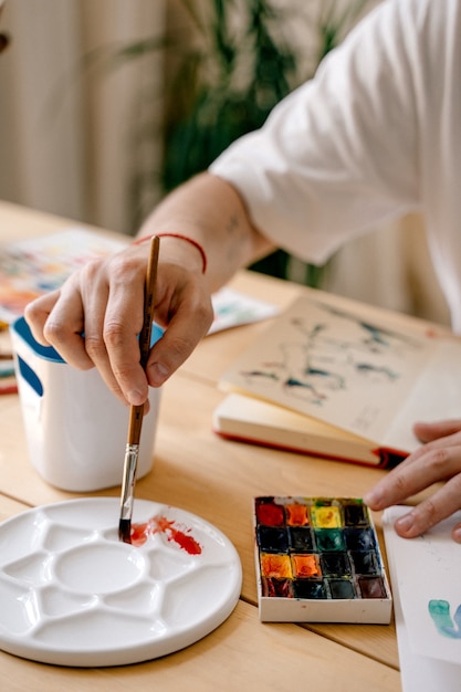 Un jeune artiste peint à l'aquarelle dans un cahier assis à une table en bois. De belles mains masculines tiennent un pinceau. Fermer. Rayons de soleil et ambiance agréable. concept artisanal. L'artiste en action