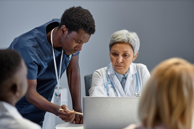 Jeune assistant confiant en uniforme bleu pointant sur l'écran d'un ordinateur portable pendant la présentation et expliquant les données en ligne à un médecin expérimenté