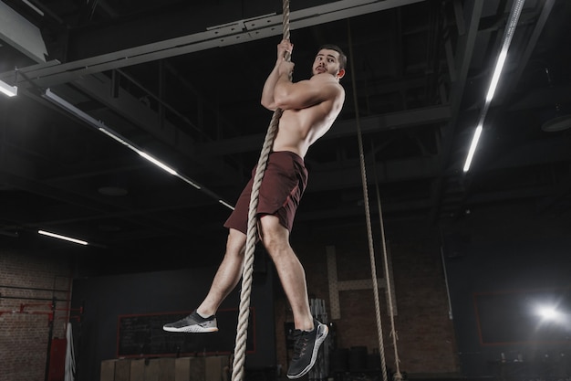 Jeune athlète crossfit escalade une corde au gymnase. Homme faisant une formation fonctionnelle. Exercices d'entraînement