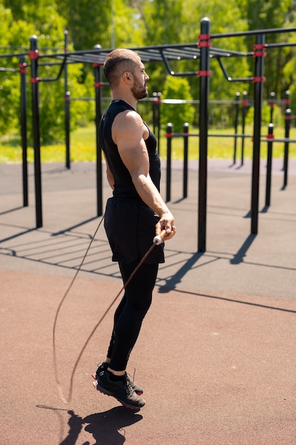 Jeune athlète musclé en survêtement sautant avec corde à sauter le matin ensoleillé pendant l'entraînement en plein air