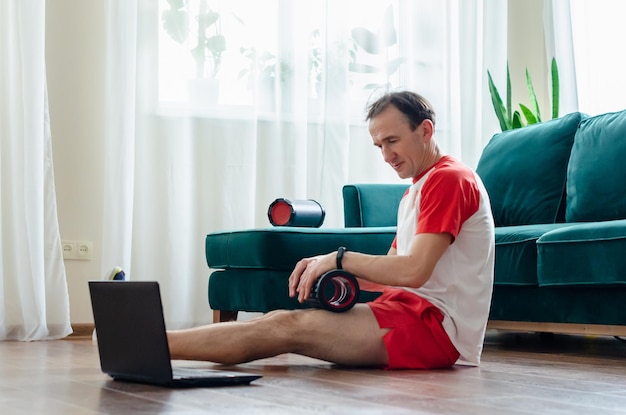 Un jeune athlète en short rouge fait des exercices sur un rouleau en mousse à l'aide d'un ordinateur portable. Travaille les muscles avec un rouleau de massage. Sports en ligne ou tutoriel vidéo suivant