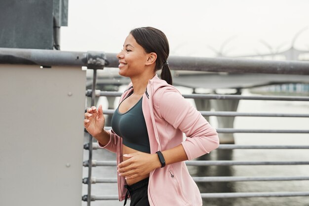 Jeune athlète souriant en vêtements de sport courant le long du pont le matin à l'extérieur