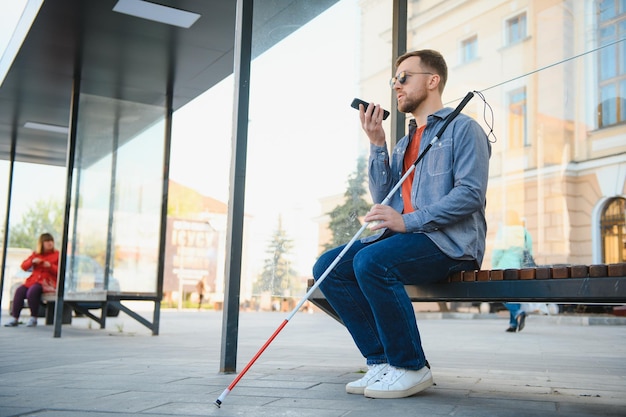 Jeune aveugle avec smartphone assis sur un banc dans le parc en ville appelant