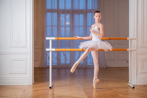 Jeune ballerine élancée dans un tutu blanc debout sur la pointe à un bar de ballet dans une belle salle blanche devant un miroir.