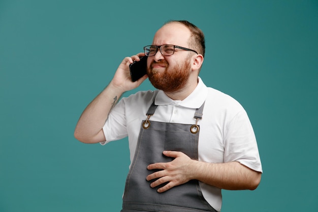 Jeune barbier irrité en uniforme et lunettes gardant la main sur le ventre regardant à côté tout en parlant au téléphone isolé sur fond bleu