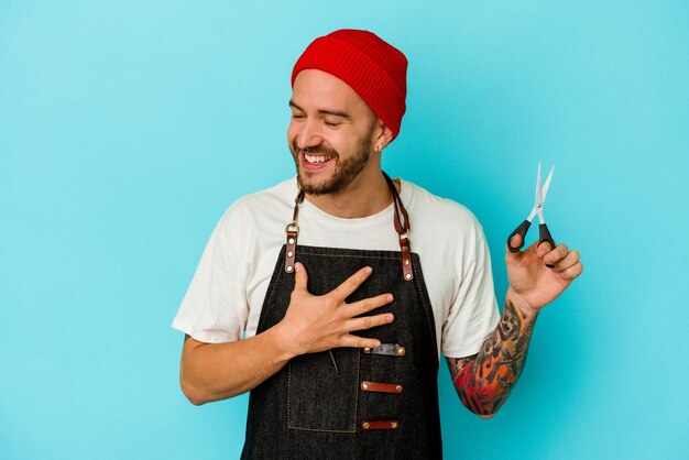 Jeune barbier tatoué isolé sur fond bleu rit fort en gardant la main sur la poitrine.