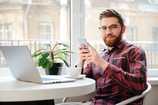 Jeune barbu à l'aide d'un téléphone portable et d'un ordinateur portable.