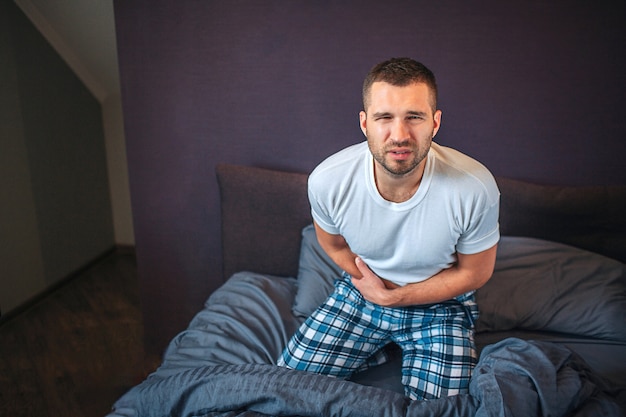 Jeune et barbu se tient à genoux sur le lit et regarde la caméra. Il se rétrécit. Guy tient la main sur la zone de l'annexe. Il ressent de la douleur. Guy souffre.