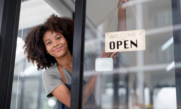 Jeune barista africaine souriante avec tablier avec panneau ouvert debout à l'entrée du magasin