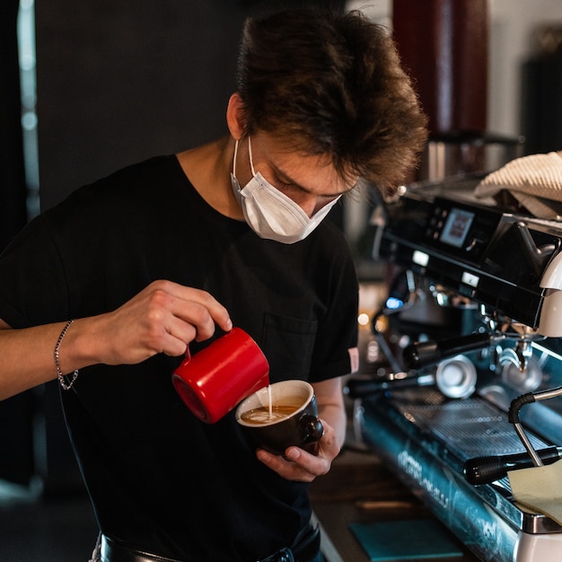 Un jeune barista avec un masque médical dans un t-shirt noir fait un délicieux café près d'une machine à café dans un café. Notion de coronavirus